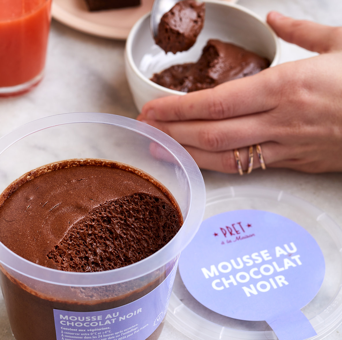 Un pot entamé de mousse au chocolat noir et une personne qui mange une cuillerée de mousse avec un verre de jus orange-fraise en arrière plan.