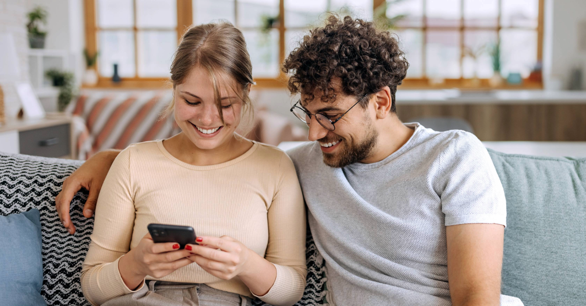 En ung kvinne og en ung mann sitter sammen på en sofa i en lys stue. Hun holder en mobiltelefon og smiler mens hun viser ham noe på skjermen. Han sitter tett inntil, ser på skjermen og smiler. Stemningen er avslappet og hyggelig.