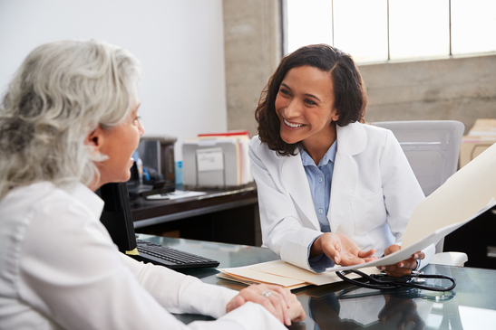 Eine ältere Frau spricht mit einer Ärztin, welche lächelt und Krankenakte bespricht.