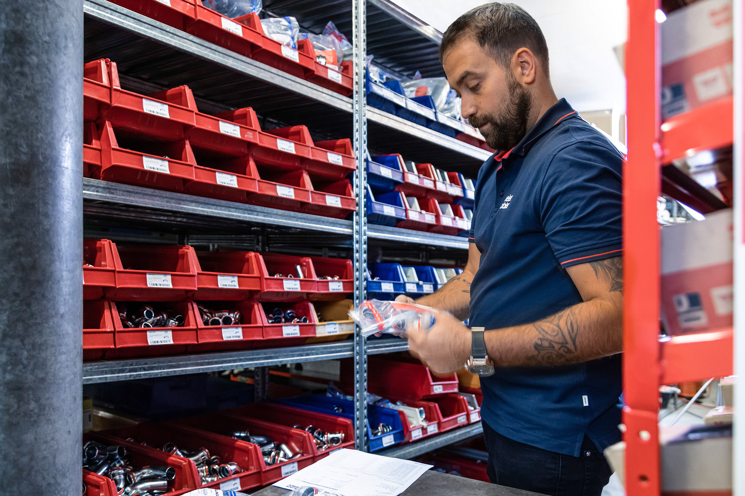 Mann in blauem Poloshirt prüft Kleinteile vor Lagerregal mit roten und blauen Behältern in Werkstatt. Mann in blauem Poloshirt prüft Kleinteile vor Lagerregal mit roten und blauen Behältern in Werkstatt.