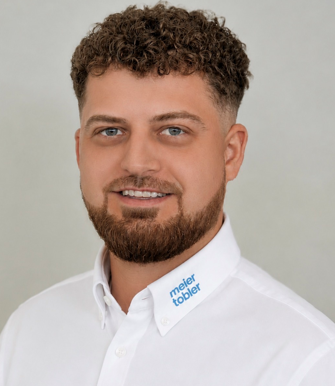 Mann mit braunen Locken und Bart lächelt in weißem Hemd mit blauem Logo vor hellem Hintergrund. Mann mit braunen Locken und Bart lächelt in weißem Hemd mit blauem Logo vor hellem Hintergrund.