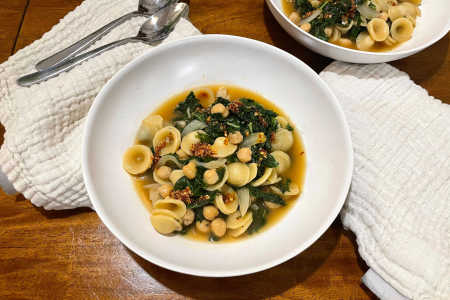 Two bowls of pasta with broth, kale and chickpeas on a wooden table with two napkins and two spoons.