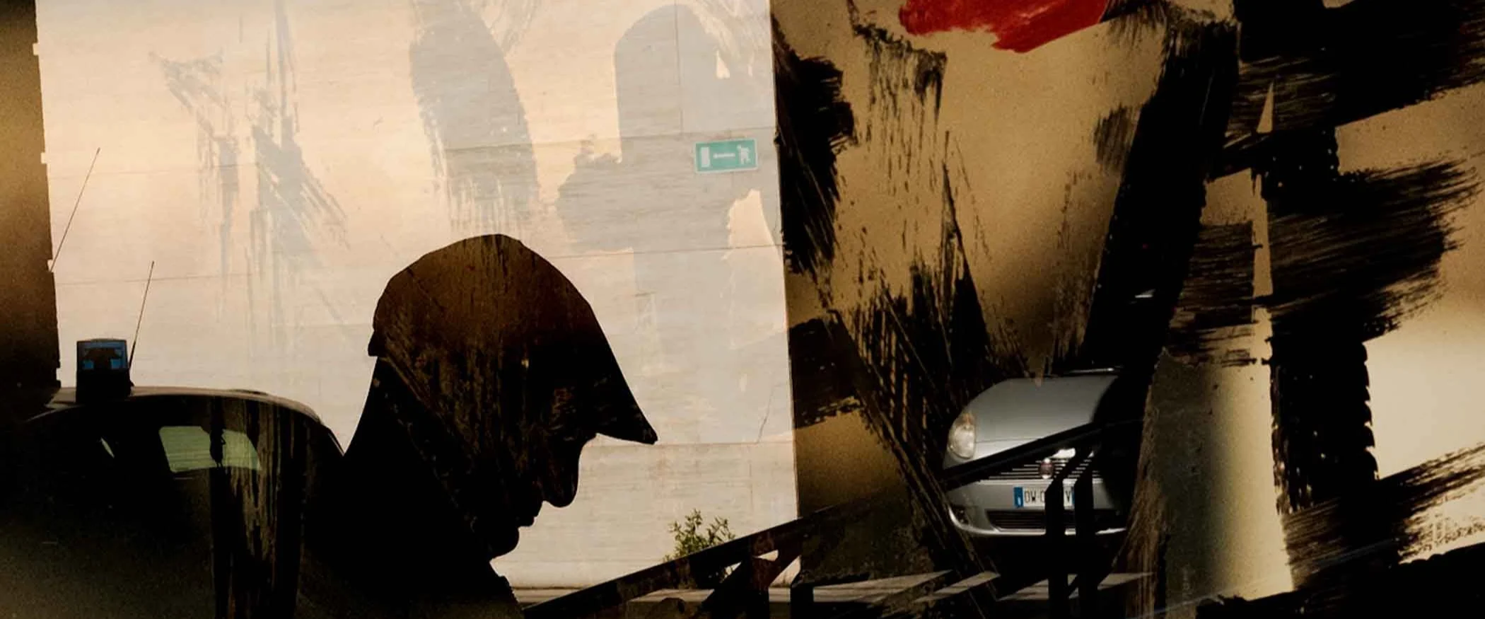 Silhouette eines Mannes mit Hut vor einer Wand mit Schatten und abstrakten roten und schwarzen Pinselstrichen. Ein Teil eines Autos ist zu sehen.