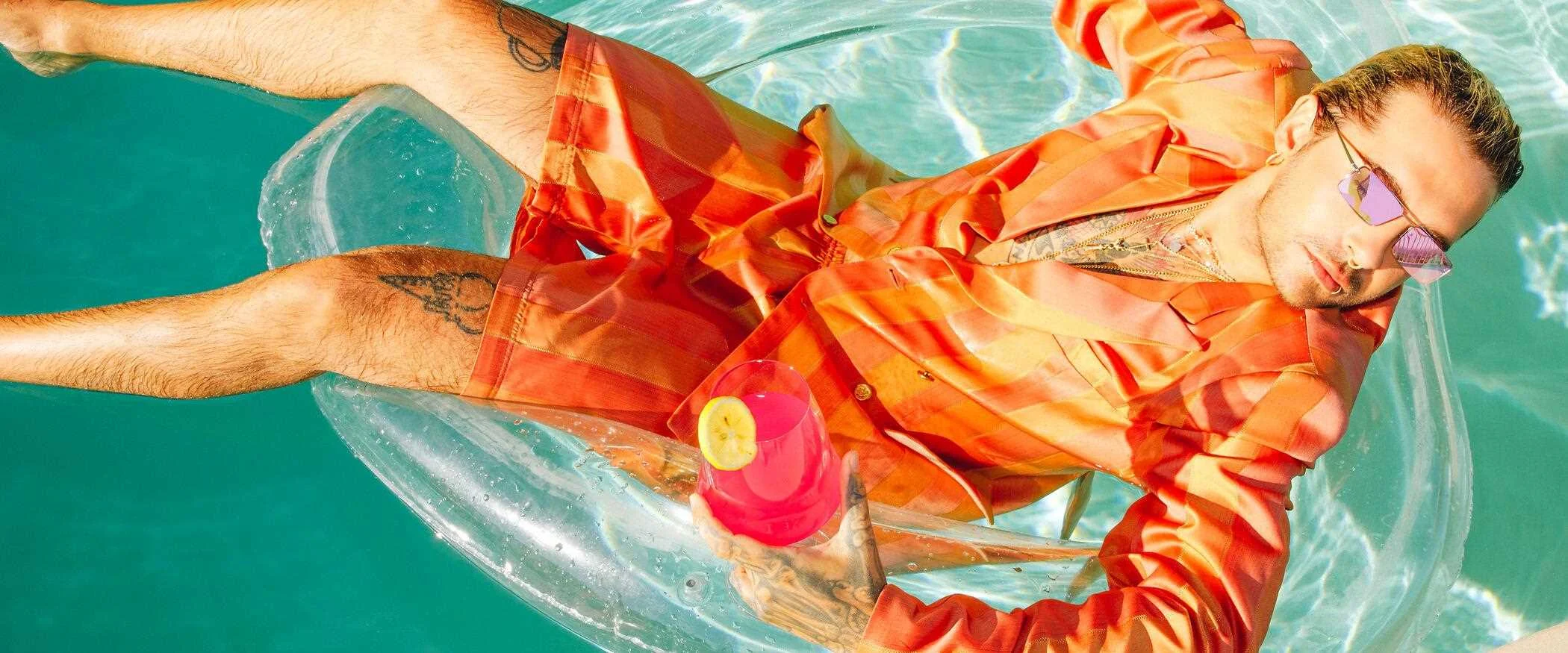 Mann in orangefarbenem Outfit und mit pinken Sonnenbrillen entspannt auf einer Pool-Luftmatratze und hält einen pinken Cocktail mit einer Zitronenscheibe.
