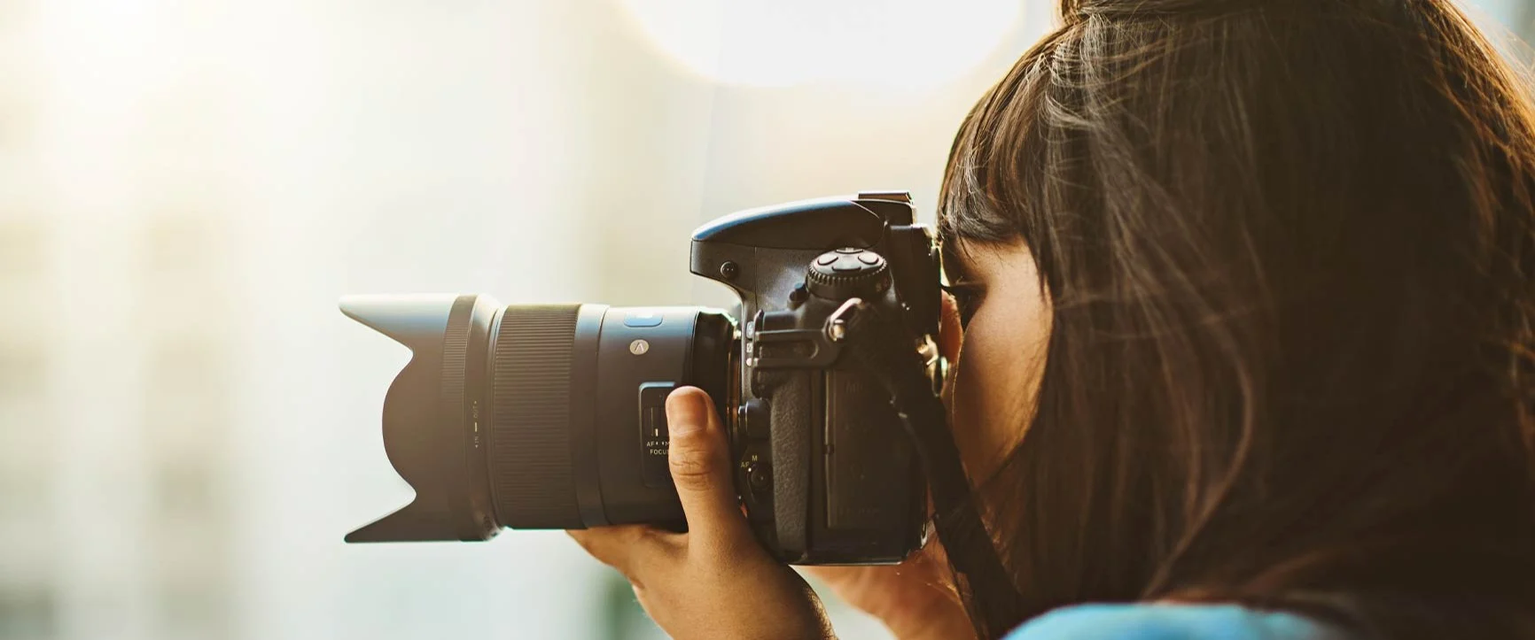 A woman with dark hair is holding a camera with a large lens and looking through the viewfinder.