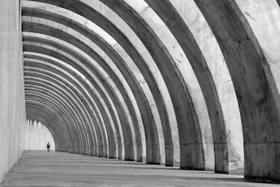 Bildaufbau Schwarz-weiß Foto mit bogenförmigen Betonstrukturen und einer Person in der Ferne.