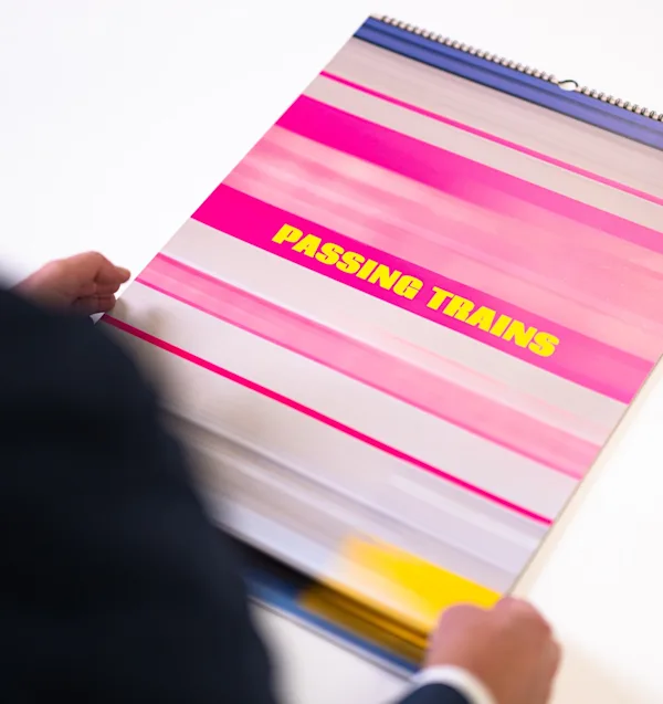 Fotografo con un calendario Una persona tiene in mano un calendario con rilegatura a spirale intitolato Passing Trains in testo giallo, con una copertina astratta a righe rosa e bianche.