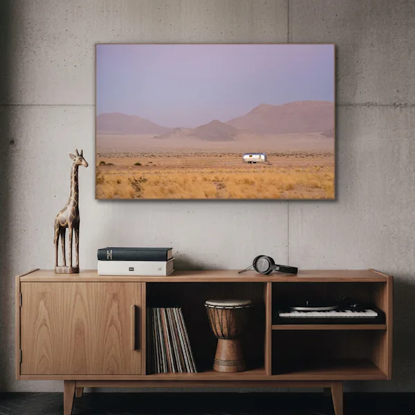 Présentation d'une décoration murale dans un intérieur moderne Un tableau de paysage encadré est accroché sur un mur en béton gris au-dessus d'un buffet en bois clair. L'image montre un vaste paysage désertique avec une caravane Airstream argentée devant de douces chaînes de montagnes sous un ciel violet. Sur le buffet se trouvent une figurine de girafe en bois, deux livres, un casque audio, un tambour et un tourne-disque avec des vinyles.