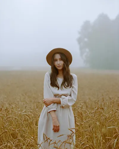Objectif & Intensité lumineuse 1 Femme ii une robe avec un chapeau se tient dans un champ par temps de brouillard. Des arbres sont visibles à l'arrière-plan.