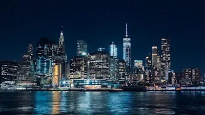 Weißabgleich und Farbtemparatur 1 Nachtszene einer beleuchteten Skyline mit hohen Gebäuden, reflektierendem Licht auf einer Wasserfläche im Vordergrund und einem klaren Sternenhimmel.