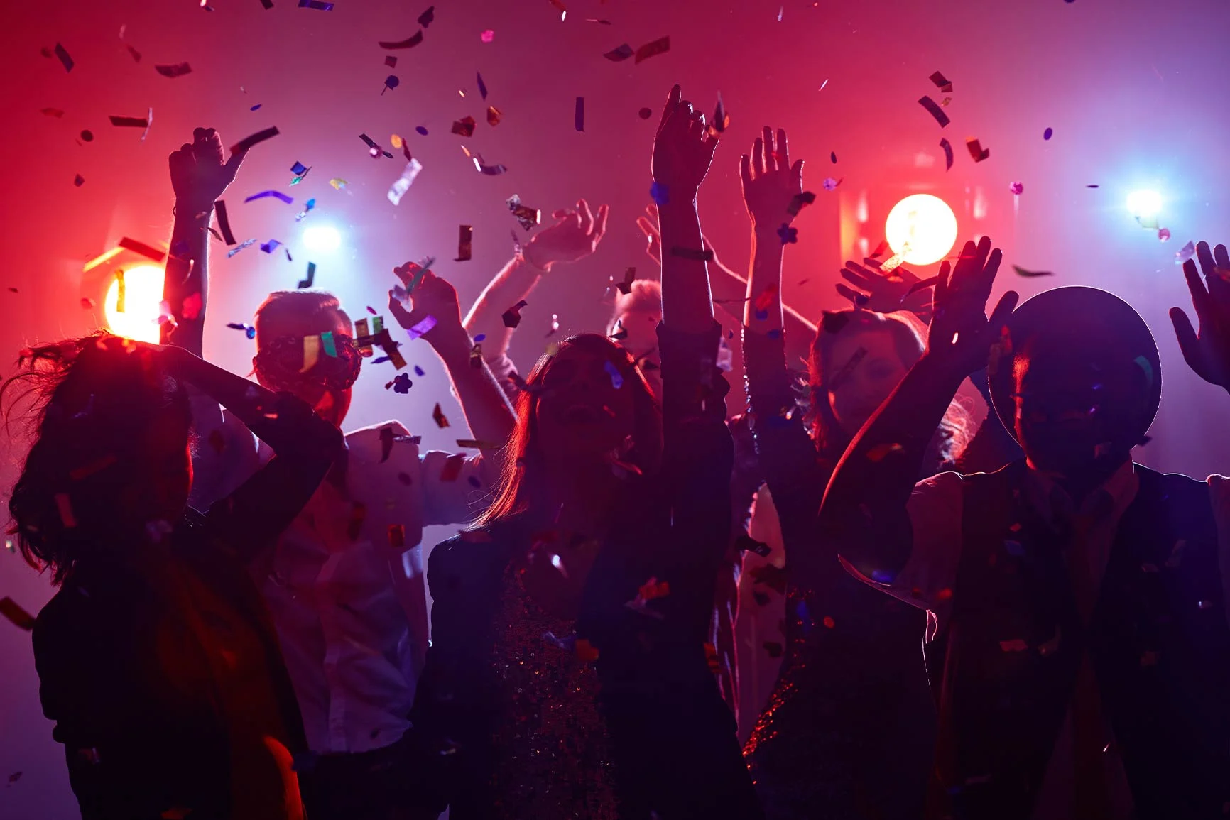 People dancing in colorful lights A group of people is dancing with their arms raised in a room filled with colorful lights and confetti.