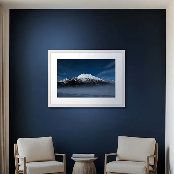 Framed Fine Art print with passe-partout in an elegant living setting A high-quality framed wall print with a wide white passe-partout (mat) is centered on a dark blue wall. The motif features a snow-capped mountain peak under a starry night sky with soft fog in the foreground. Two light-colored armchairs and a small wooden table beneath the picture emphasize the calm and sophisticated interior.