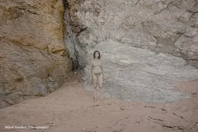 Circulation(s) Festival 2023 Person stands with sand-covered body in front of a rocky wall on a beach with light-colored sand.
