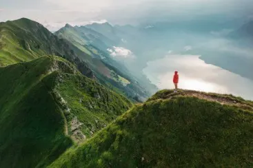 Person on mountain peak Person in red coat standing on green mountain peak overlooking mountain range and lake in fog.
