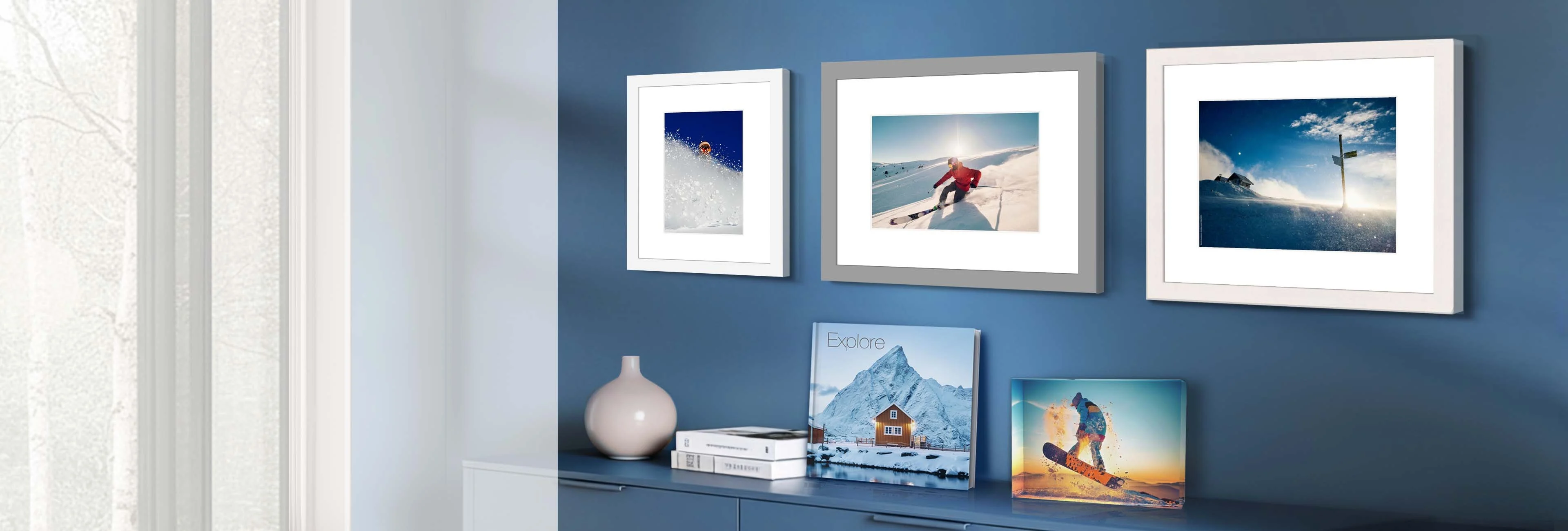A perspective view of a modern interior. Three framed winter sports motifs with wide white passe-partouts hang on a blue wall in different frame colors (white and gray). On the sideboard below, a square photo book titled "Explore" (featuring a mountain hut) and a transparent acrylic block with a snowboarder motif are decoratively placed.