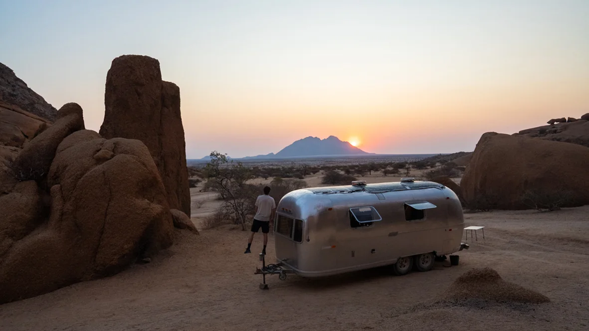 Thylacine en Namibie – Rencontre avec la nature Un cliché atmosphérique dans un paysage rocheux de Namibie au crépuscule. Au centre se dresse un arbre à l'écorce blanche frappante et aux branches largement ramifiées, presque dénudées. Derrière le tronc, un homme vêtu de sombre se tient debout, sa silhouette étant partiellement masquée par les branches. L'environnement se compose de formations rocheuses sombres et accidentées sous un ciel de soirée violet pâle.