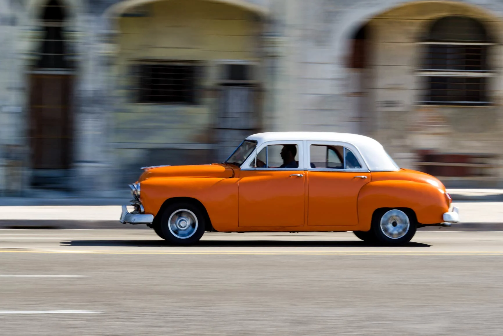 Coche clásico naranja con techo blanco pasa por delante de un edificio con arcos y ventanas. Coche clásico naranja con techo blanco pasa por delante de un edificio con arcos y ventanas.