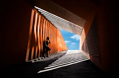 Die richtige Belichtung messen und einstellen 1 Eine Person steht in einem orangefarbenen Gang mit Lamellen, die Schatten werfen. Am Ende des Gangs ist ein heller blauer Himmel mit weißen Wolken zu sehen.