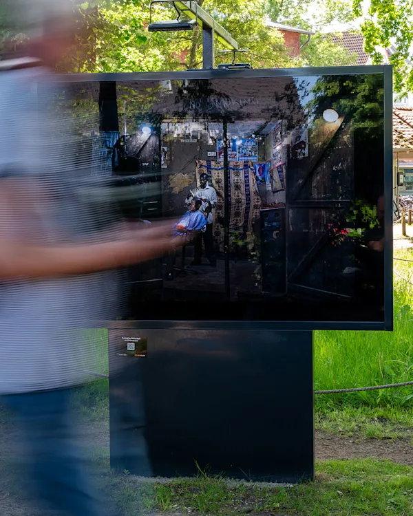 WhiteWall Outdoor Exhibition on ChromaLuxe An outdoor display board showing a photo of a barber cutting hair in a rustic shop, with a blurred person walking past.