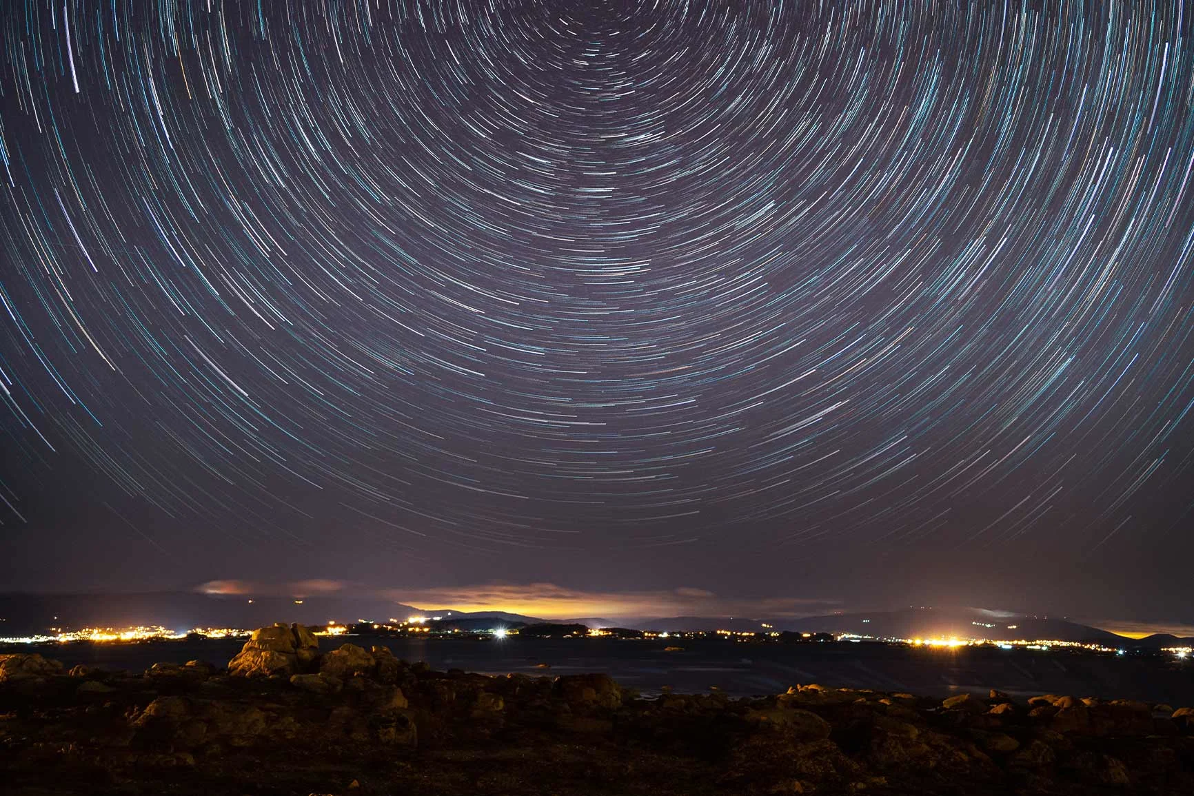 Cielo estrellado con trazas circulares de estrellas sobre un paisaje oscuro con luces en el horizonte. Cielo estrellado con trazas circulares de estrellas sobre un paisaje oscuro con luces en el horizonte.