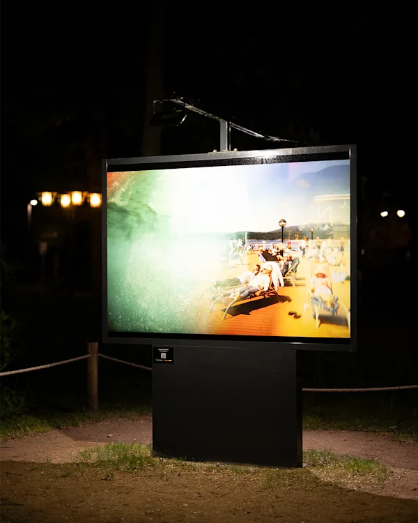 Sarah Palmer Outdoor-Exhibition 'Wish You Were Here' produced by WhiteWall An illuminated outdoor display at night showing a photo of people relaxing on sun loungers on a cruise ship deck.