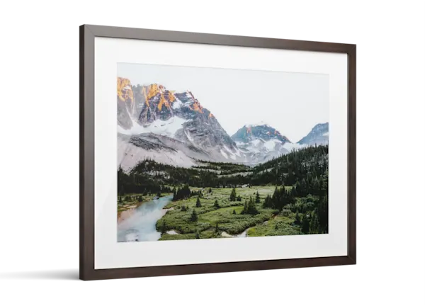 Framed print of a mountain landscape with snow-capped peaks, green valley, evergreen trees, and a turquoise river.