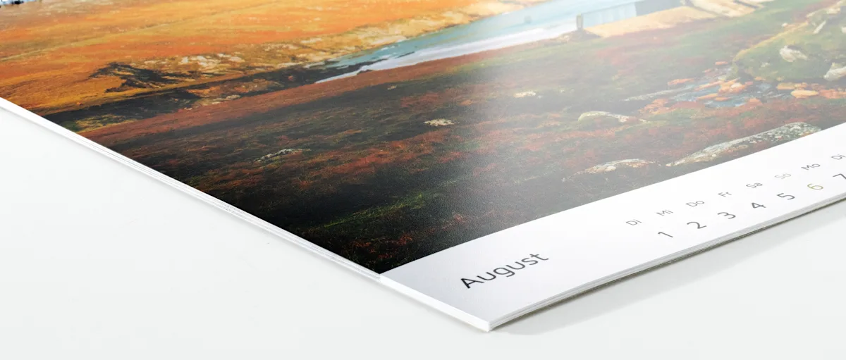 Fotowandkalender Inkjet glänzend Nahaufnahme einer Kalenderseite, die August zeigt, mit einem darüber sichtbaren Teilausschnitt einer Landschaftsaufnahme in Orange- und Dunkeltönen