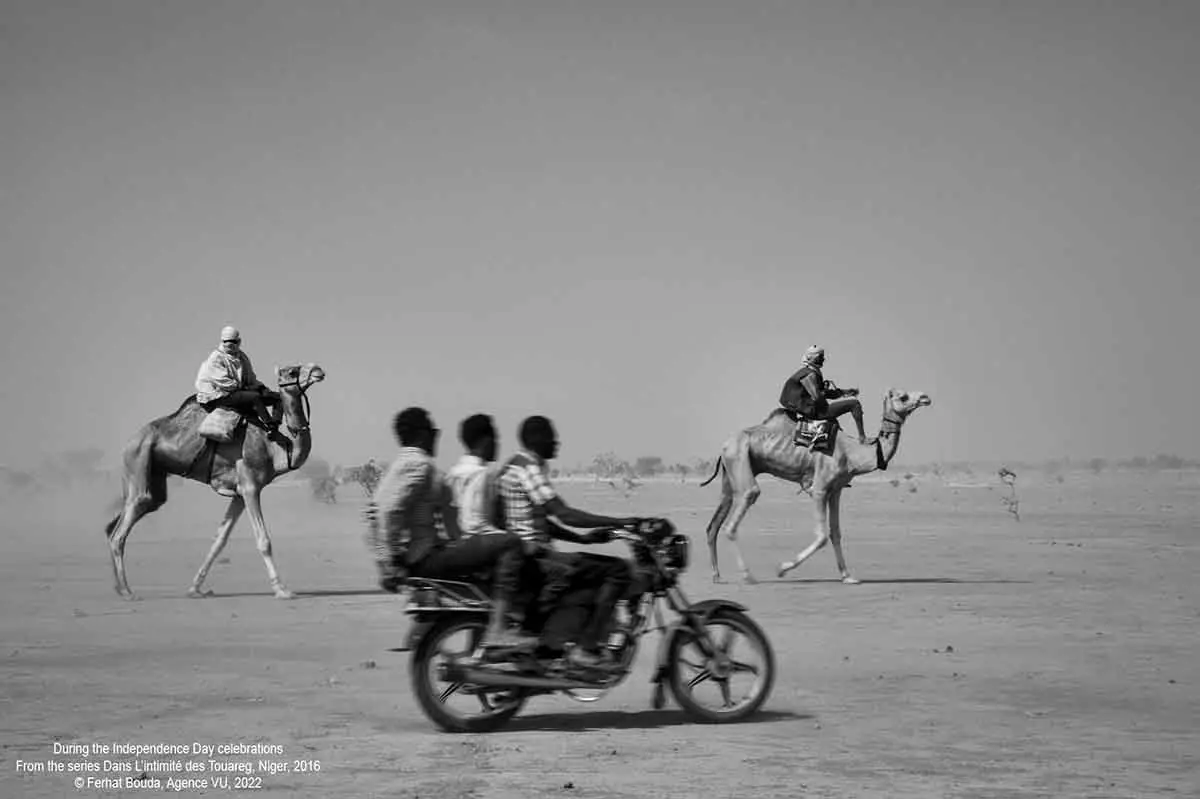 three men riding a moped next to camel riders in a desert landscape - photo by Ferhat Bouda.