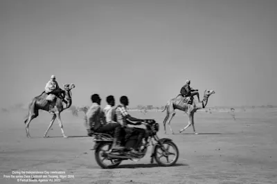 Ferhat Bouda - Photographs from North Africa Three men riding a moped next to camel riders in a desert landscape.