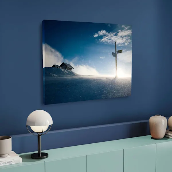 Canvas print with winter landscape in a modern interior A large-format canvas print hangs on a dark blue wall above a mint-green sideboard. The motif depicts a snowy mountain hut and a signpost in the bright backlight of the winter sun. A minimalist globe lamp and decorative vases on the sideboard emphasize the modern ambiance.