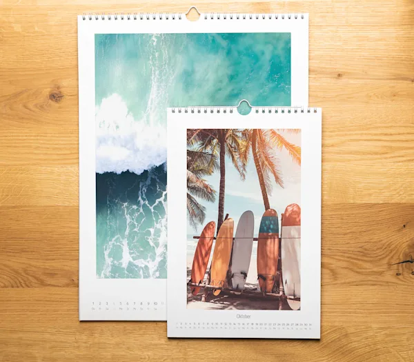 Calendari a tema viaggio Due calendari da parete con rilegatura a spirale su una superficie in legno, raffiguranti onde oceaniche e tavole da surf sotto palme per il mese di ottobre.