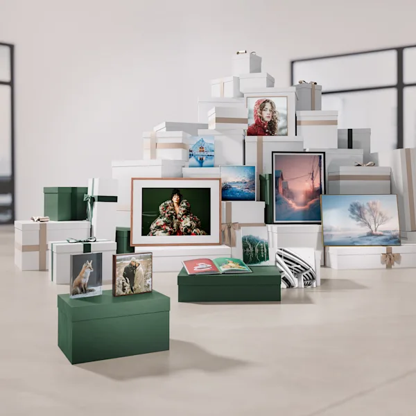 Bestellfristen für Fotogeschenke Eine große Ansammlung von weißen und dunkelgrünen Geschenkboxen, die mit beigefarbenen oder grünen Bändern verschnürt sind, ist in der Mitte eines hellen Raumes aufgebaut. Die Boxen bilden eine Pyramide oder einen Stapel. Verschiedene gerahmte Kunstdrucke und Fotos sind auf den Boxen platziert oder lehnen daran. Die Bilder zeigen unterschiedliche Motive, hauptsächlich Natur- oder Winterlandschaften, aber auch Porträts und abstraktere Darstellungen. Im Vordergrund stehen zwei dunkelgrüne rechteckige Boxen. Darauf liegen kleinere Bilder, darunter eines mit einem Fuchs und eines mit einem Paar, sowie ein aufgeschlagenes kleines Buch oder Katalog mit rot-grünem Einband. Der Boden ist hellbeige und die Wände sind weiß. Im Hintergrund sind durch große Fenster oder Türen schwache Umrisse eines Innenraums zu erkennen. Die Szene vermittelt den Eindruck einer Präsentation von Produktvielfalt, vielleicht für die Feiertage.