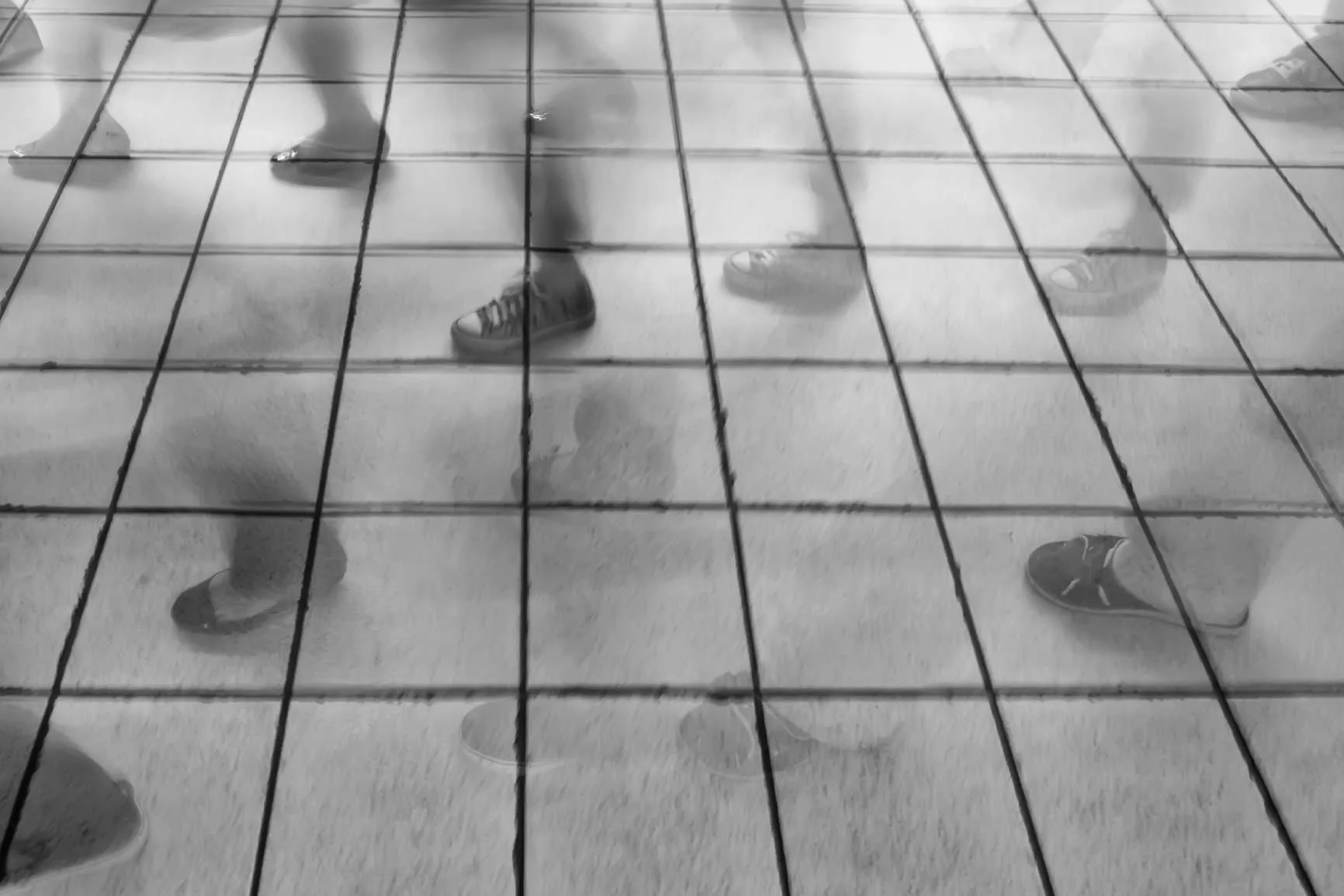 Varias piernas y pies borrosos con sandalias y zapatos caminan sobre un suelo de baldosas con juntas visibles. Varias piernas y pies borrosos con sandalias y zapatos caminan sobre un suelo de baldosas con juntas visibles.
