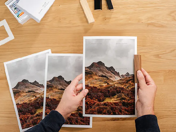 WhiteWall Produktmuster Hände, die gedruckte Landschaftsfotos einer herbstlichen Berglandschaft auf einem Holzschreibtisch anordnen, mit schroffen Gipfeln und orangefarbenem Laub