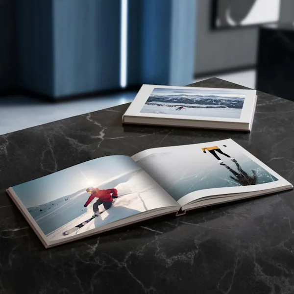 Open coffee table book on a marble table A high-quality, square-format photo book lies open on a dark marble surface. The pages display dynamic winter sports photography: a skier in deep powder and a skater on a reflective ice surface. In the background, another closed photo book and a modern interior are visible.