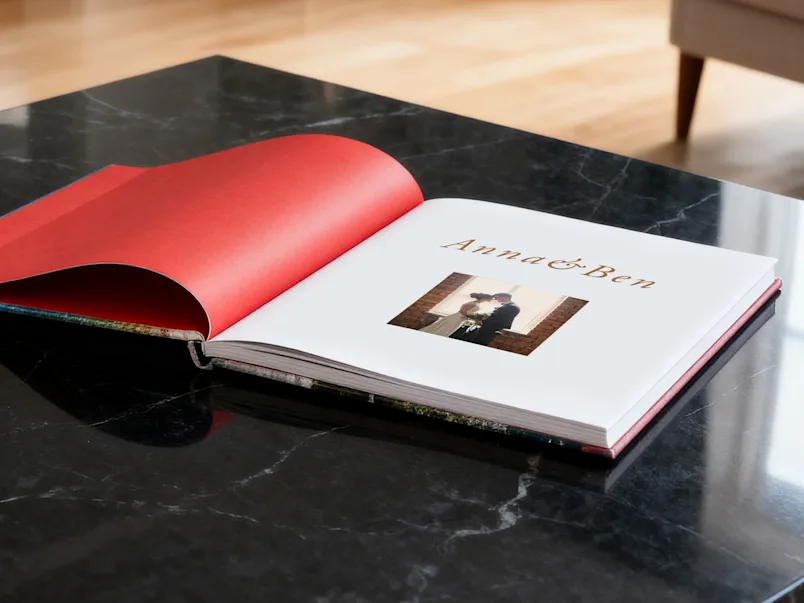 SEA / SEO LP | Wedding Photo Book | Product Selector Section | Media #3 Álbum de fotos de boda abierto titulado «Anna & Ben» sobre una mesa de mármol negro, que muestra las guardas rojas y una foto de la pareja.