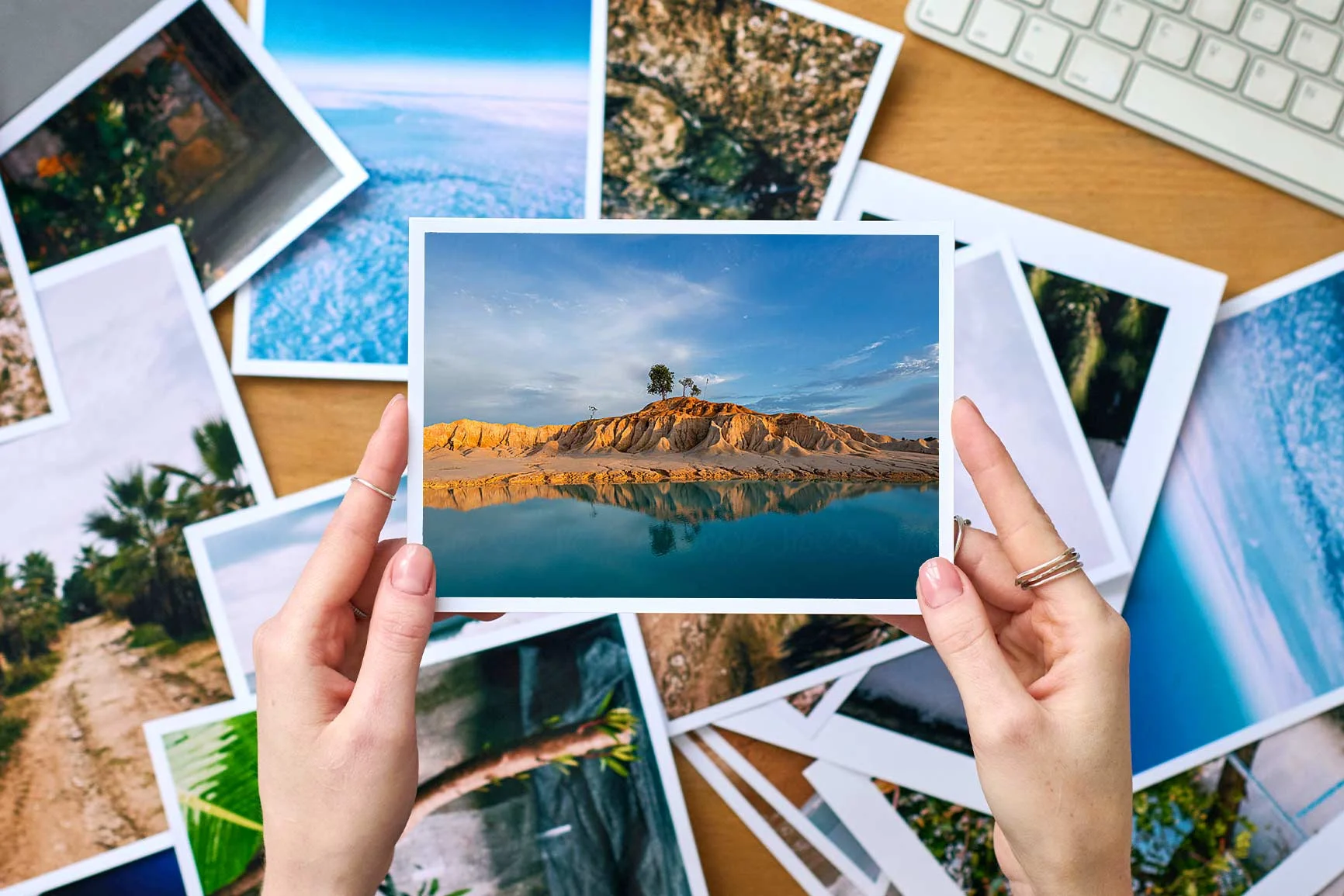 Le mani reggono una fotografia di un'isola con un albero e il suo riflesso nell'acqua, circondata da altre fotografie. Le mani reggono una fotografia di un'isola con un albero e il suo riflesso nell'acqua, circondata da altre fotografie.