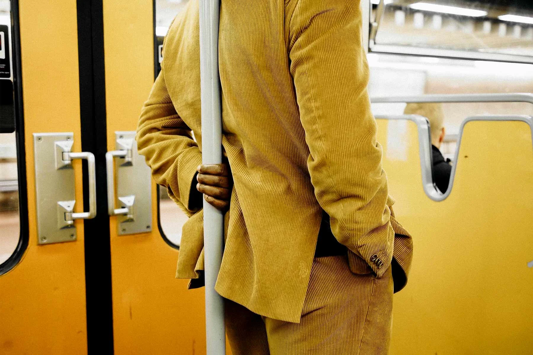 Una persona con traje beige está de pie en un compartimento de tren amarillo y sostiene una barra con la mano izquierda. Una persona con traje beige está de pie en un compartimento de tren amarillo y sostiene una barra con la mano izquierda.