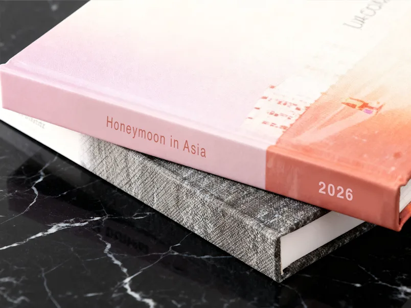 SEA / SEO LP | Wedding Photo Book | Product Selector Section | Media #4 Dos fotolibros apilados sobre una superficie de mármol; el de arriba es rosa y lleva impreso en el lomo «Luna de miel en Asia 2026».