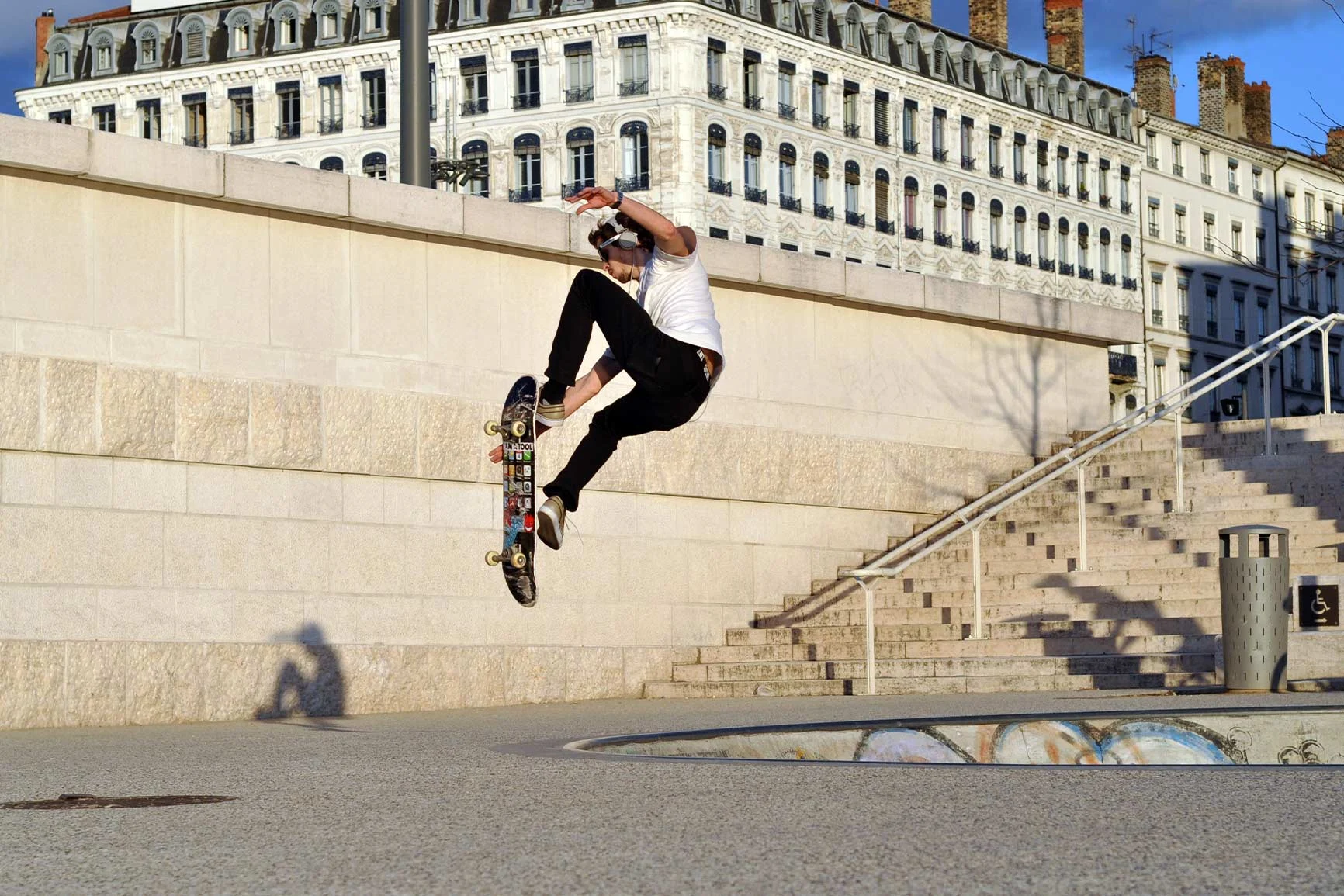 Skateboarder jumps in front of a staircase A skateboarder jumps in front of a set of stairs in an urban setting with historic buildings in the background.