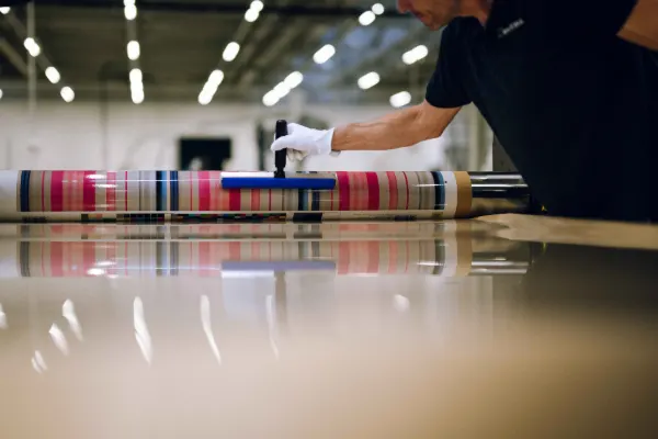 Professional photo print production WhiteWall lab In a modern production facility, a technician wearing a white protective glove carefully processes a large, glossy photo print. The print features a colorful pattern of vertical pink, beige, and blue stripes and is wrapped over a cylindrical metal roller. The worker uses a hand squeegee tool to smoothly mount the image, which reflects clearly on the polished work surface in the foreground. In the soft-focus background, rows of industrial ceiling lights illuminate the manufacturing hall.