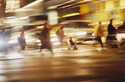 Straßenfotografie Verschwommene Fußgänger überqueren nachts eine belebte, beleuchtete Straße mit Autos im Hintergrund.