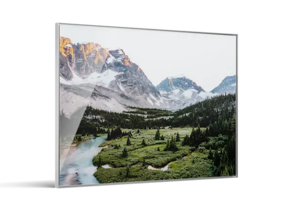 Framed landscape print showing snow-capped mountains, green valley with evergreen trees, and a winding turquoise river below.