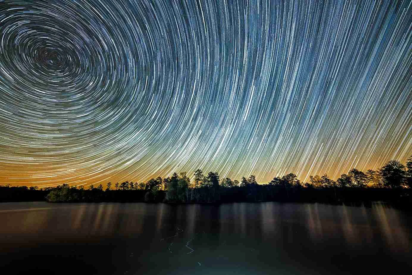 Cielo estrellado con trazas circulares de estrellas sobre un lago y una hilera de árboles en el horizonte al atardecer. Cielo estrellado con trazas circulares de estrellas sobre un lago y una hilera de árboles en el horizonte al atardecer.