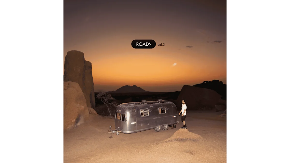 Thylacine en Namibie – Studio mobile dans le paysage désertique Une vue large au crépuscule dans un paysage de désert rocheux en Namibie. Au centre se trouve une caravane Airstream argentée et brillante, servant de studio d'enregistrement mobile. Un homme se tient à droite du véhicule sur une petite butte de sable et regarde vers l'appareil photo. L'environnement est marqué par un sol sablonneux et des formations rocheuses sombres et monumentales qui se détachent sur un ciel du soir orange intense.