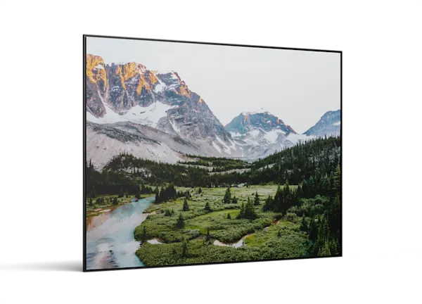 A framed landscape photo showing snow-capped mountains, a green valley with evergreen trees, and a winding blue river below.