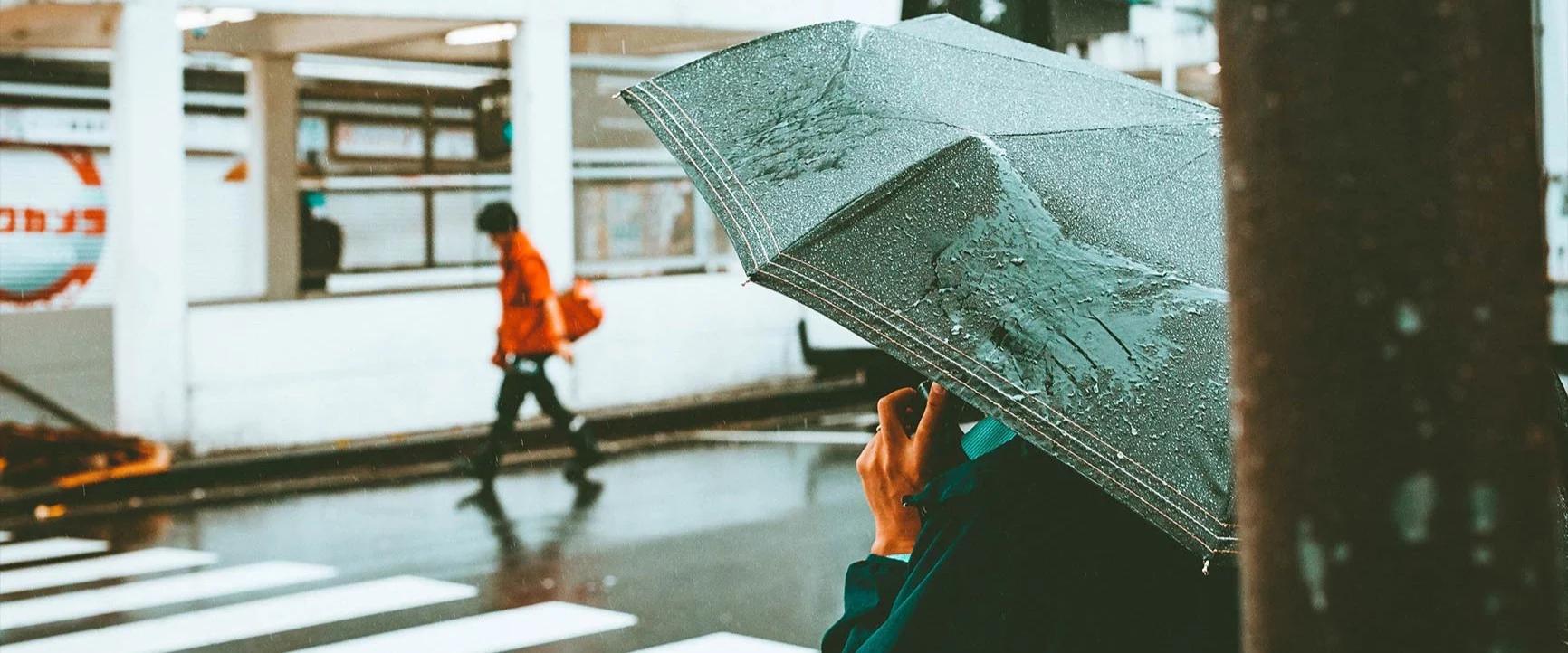 A person holding an umbrella in front of a crosswalk in the rain, with another person in the background.