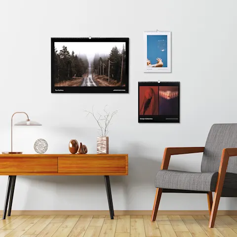 Visualizzazione dei calendari Tre calendari fotografici esposti su una parete bianca sopra una credenza in legno in stile moderno di metà secolo e una poltrona grigia, in un elegante soggiorno.