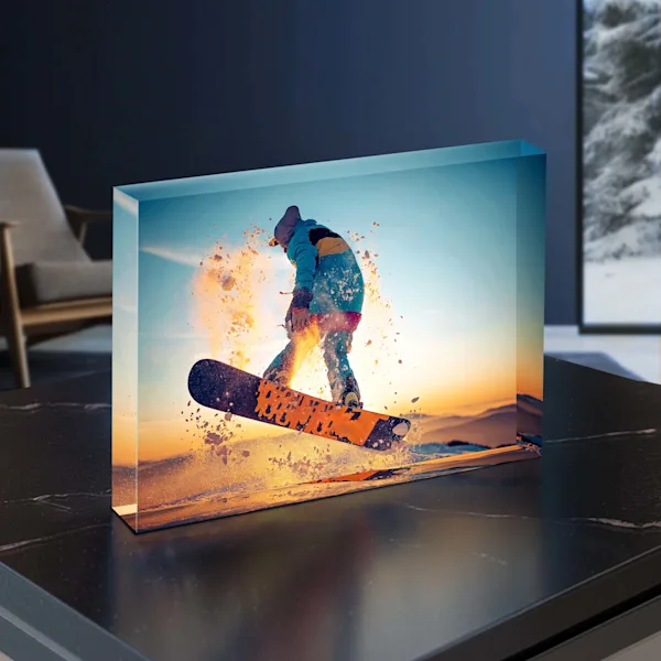 Personalized acrylic block with winter sports motif A free-standing, transparent acrylic block sits on a dark marble surface. The block features a vibrant photo of a snowboarder jumping during sunset. The thick acrylic layer gives the image impressive depth and brilliance. In the blurred background, a modern living room interior is visible.
