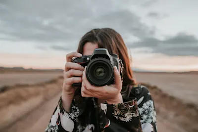 Choisir le bon appareil photo 1 Une personne aux cheveux noirs et aux vêtements à motifs tient un appareil photo devant son visage et prend des photos dans un vaste environnement paysager, sous un ciel nuageux.